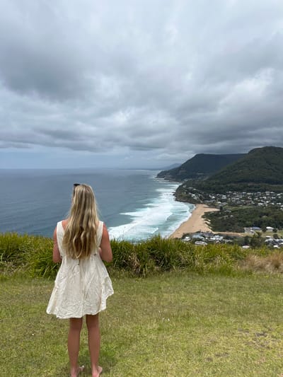 A woman stands on a grassy cliff, gazing at a beach and ocean view. She wears a light-colored dress, and her long blonde hair flows down her back. The ocean's waves crash against the sandy shore, with a line of buildings nestled along the beach. A mountain covered in trees forms a backdrop, and the sky is overcast with grey clouds.