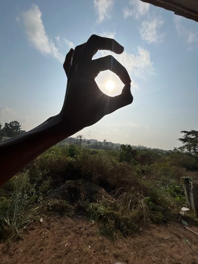 The image captures an outdoor scene in Debasco, Nigeria, under bright daytime conditions. In the immediate foreground, a human hand is silhouetted against a light blue sky, forming an "OK" gesture with the thumb and index finger to perfectly encircle the sun. The sun emits bright rays of light, causing a prominent lens flare effect. The sky above is largely clear, with scattered white cumulus clouds primarily in the upper left quadrant.
The lower half of the frame depicts a landscape composed of reddish-brown soil in the foreground, partially covered by sparse green and dry brown vegetation. Towards the mid-ground, the vegetation becomes denser, forming a thick layer of wild grasses, shrubs, and small trees. A small, dark mound of material, possibly soil or debris, is visible on the ground to the left. In the distant background, a hazy horizon reveals more extensive tree cover, including discernible palm trees, and the faint outlines of residential or urban structures, suggesting a developed area beyond the immediate wilderness. A fragmented, weathered structure, possibly a wall, is partially visible on the far right edge of the foreground, with incidental white plastic bags or debris resting against it. The overall lighting is characterized by strong backlighting from the sun, rendering the foreground elements largely in silhouette.