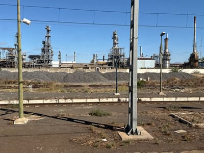 The image depicts an industrial landscape in Sidi Kacem, Morocco, characterized by a large refinery complex viewed from a railway area. In the immediate foreground, a dark, dusty ground with sparse dry vegetation and debris is visible. Two vertical poles, likely part of railway infrastructure, stand prominently: a thinner greenish pole on the left with a white rectangular sensor-like object near its top, and a thicker grey metallic lattice pole on the right. Overhead catenary wires span horizontally across the upper frame. Below the poles, a concrete railway platform edge is visible, bordering at least two parallel railway tracks.

In the midground, behind the tracks, large, irregular mounds of dark, granular material are piled across the scene, separating the railway from the main industrial facility. Scattered debris and smaller poles are interspersed among these piles.

The background is dominated by an extensive industrial complex, consistent with a refinery. Numerous tall, grey metallic structures, including distillation columns, interconnected piping networks, and smokestacks, are present. Some structures exhibit signs of weathering and rust. A light-colored rectangular building is visible in the center-right of the complex. Sparse vegetation, including a few palm trees, borders the industrial site. The sky is clear and blue, and sharp shadows indicate strong sunlight. No individuals are present in the scene. In the far distance on the right, low hills are visible.