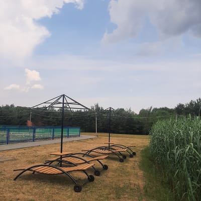 A row of empty wooden lounge chairs with metal frames are placed on a dry, grassy field. Two umbrella frames stand behind the chairs. In the background, a blue fence encloses a green area, possibly a sports court. Beyond the fence is a dense line of trees. The sky is partly cloudy with a mix of white and gray clouds against a light blue backdrop. The scene is located in Dubrivka, Ukraine.