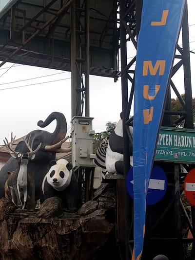 A whimsical animal display features an elephant, two deer, a panda, and a zebra, arranged atop a faux rocky terrain. An electrical box stands near the elephant. A blue banner with vertical yellow lettering reads "LMUW," and a green sign mentioning "KAPTEN HARUN" sits behind. The scene is framed by a metal structure, possibly a utility pole, and a cloudy sky provides the backdrop.