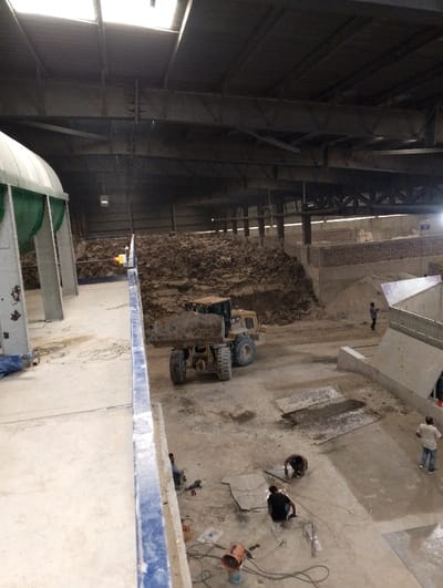 The image shows an industrial setting, likely a construction or manufacturing site, located at 22.693 latitude and 90.601 longitude in Pangasia, Bangladesh. The scene is captured from an elevated perspective looking down upon a large, open space. A sizable pile of dirt or aggregate material occupies the central area, with a yellow CAT-brand dump truck parked to the right of it. Several workers are scattered around the site, engaged in various tasks, such as welding metal sheets, inspecting equipment, and possibly preparing for concrete pouring. The workers appear to be wearing casual clothing. The area is dimly lit and dusty, with exposed steel beams and rafters supporting the roof. The scene is characterized by the presence of industrial machinery, construction materials, and the activities of the workers.