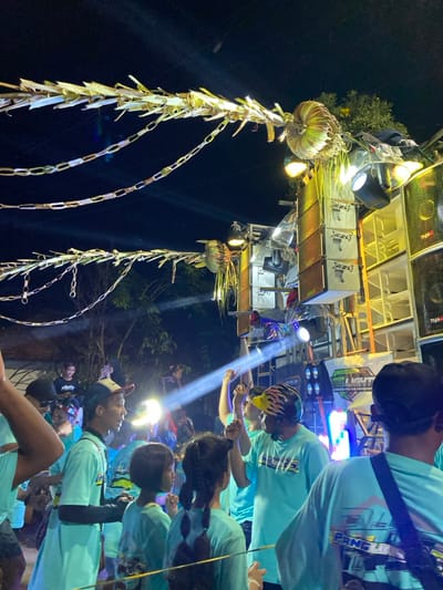 At a nighttime outdoor event in Kedungbanteng, Indonesia, a crowd of individuals is gathered under an array of decorative and technical elements. Overhead, multiple strands of pale, possibly organic, chain-like decorations are strung across the frame against a dark sky, illuminated by ambient light. These decorations feature larger, spiky, rounded elements attached to support structures, also appearing to be crafted from organic materials.

To the right, a significant portion of a large, multi-tiered sound and lighting rig is visible. This rig comprises numerous stacked white or light grey speaker cabinets, many bearing the "RZL" logo, alongside other speakers and large subwoofers. Integrated stage lights emit bright beams, with one prominent white light beam cutting horizontally across the midground. A banner displaying "JS LIGHTING" and additional text is affixed to the lower section of the rig.

In the foreground and midground, a diverse group of people, including adults and children, are present. Many individuals are uniformly dressed in light blue or turquoise t-shirts, some featuring graphic designs. Several people have their hands raised, indicating engagement with the event. A man wearing a yellow and black patterned cap stands in the midground, facing right and appearing to interact with someone off-camera. Two young girls with braided hair, also in light blue shirts, are positioned in the midground. In the far background, a person is seen using a smartphone, illuminated by its screen, likely recording or photographing the event. The scene is lit predominantly by the stage lights, casting a strong glow on the participants and equipment.
