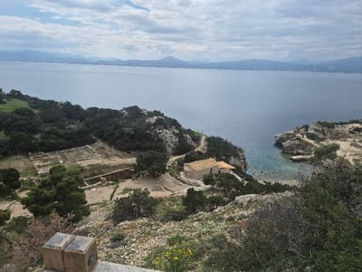 An elevated, wide-angle photograph depicts a coastal landscape in the Municipal Unit of Loutraki - Perachora, Greece, under an overcast sky.
In the immediate foreground, a weathered wooden post occupies the lower left corner, set against rocky, overgrown terrain dotted with sparse green shrubs and small yellow and white flowers.
The mid-ground features a significant archaeological site to the left, characterized by excavated stone foundations and low walls laid out on a beige, earthen surface, interspersed with scattered trees and dirt pathways. To the right, a small, light-colored building with an orange-tiled gabled roof is positioned on a rugged promontory, overlooking a secluded, clear turquoise cove. A winding dirt path leads towards this building from the upper terrain. Below the building, near the water's edge of the cove, a blue and white Greek flag is visible. Dense dark green foliage, primarily shrubs and trees, covers much of the hilly terrain separating the foreground from the sea and surrounding the structures.
The background is dominated by a vast expanse of calm, deep blue-grey water, likely a gulf or bay, with subtle reflections of light on its surface. Beyond the water, a hazy chain of blue-grey mountains stretches along the distant horizon. The sky is largely overcast, displaying various shades of grey and white clouds. No human figures or discernible animal life are present in the scene.