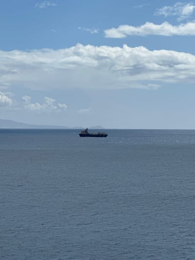 The image captures a wide view of the open sea off the coast of San Sebastián de la Gomera, Spain. A dark-hulled cargo or container ship is positioned horizontally across the mid-ground, appearing relatively distant. The vessel features a superstructure towards its stern (right side) and is situated on a moderately rippled, dark blue-grey ocean surface, which fills the lower two-thirds of the frame. In the distant upper-left quadrant, a faint, low-lying landmass is visible on the horizon, partially obscured by atmospheric haze. The upper third of the scene consists of a light blue sky with scattered white cumulus clouds, some larger and more defined towards the top, and smaller, greyer formations nearer the horizon. No individuals or specific actions are discernible within the frame.