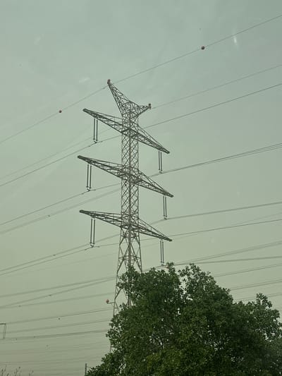 A low-angle shot captures an electrical transmission tower with three distinct support structures, set against a pale, overcast sky. The metal tower is intricately crisscrossed and connected to high-voltage power lines that stretch across the frame. Orange marker balls are attached to the lines. A portion of a tree with dense green foliage occupies the right side of the image. The location is Lod, Israel, deduced from the presence of high-voltage towers typical of infrastructure found there. No people are visible.