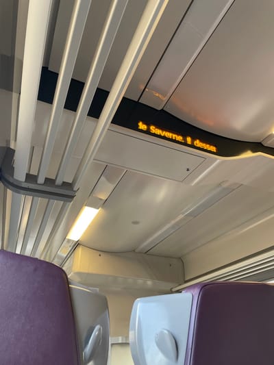 The image depicts the interior of a modern passenger train carriage, observed from a seated position looking forward and slightly upward, located in Hochfelden, France. The ceiling is primarily white and light grey, featuring modular panels and integrated linear strip lighting. A white, slatted overhead luggage rack is visible on the upper left side of the frame. Centrally positioned above the seats is an amber LED digital display screen showing the text "le Saverne. Il desser" (meaning "to Saverne. It serves/stops"). In the foreground, the upper backs of two passenger seats are visible, composed of light grey plastic shells with integrated handgrips and partially visible purple fabric upholstery. No individuals are present within the observable frame.