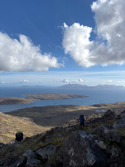 The image shows a landscape scene with a prominent rocky outcrop in the foreground, a wide expanse of water with islands in the middle distance, and a sky filled with clouds. Two figures are visible, seemingly hiking on the edge of the rocky outcrop. The environment suggests a mountainous or coastal terrain, likely a part of the Isle of Skye in the United Kingdom. The weather appears clear with sunny conditions allowing good visibility across the scene.