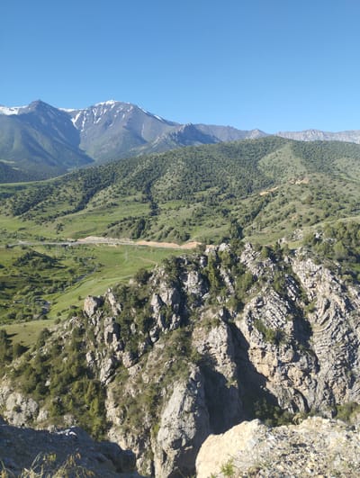 The image depicts a panoramic view of a mountainous, arid landscape. The foreground features rugged, rocky terrain with sparse vegetation. In the midground, rolling green hills are covered in dense shrubs and trees, with a road winding through the landscape, accompanied by utility poles. A small stream is visible in the valley. The background is dominated by majestic, snow-capped mountains under a clear blue sky. The scene suggests an outdoor environment with natural beauty and evidence of human infrastructure. The location is identified as Myk, Uzbekistan.