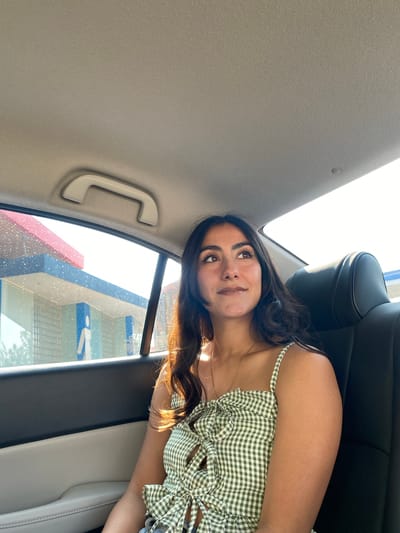 A medium shot captures a young woman in the back seat of a car. She is wearing a green and white gingham top with spaghetti straps. Her long, dark hair cascades around her shoulders. Her gaze is directed upwards and slightly to the left. The car's interior is visible, including the seat, headrest, and ceiling. Outside the window, abstract shapes and colors are visible. Based on the color scheme and shape of the building it looks like the building is a Wal-Mart. Santa Ana Xalmimilulco, Mexico, is not visible within the image, it is not possible to determine if the image was taken there based on the available data.