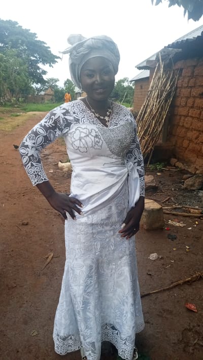 A woman stands in a dirt yard in what appears to be a rural setting. She is wearing a white, long-sleeved dress with intricate lace detailing and a white head wrap. Her hands are on her hips, and she is smiling. In the background, there is a mud brick building, a pile of wooden poles, and trees. A chicken is visible in the background. Based on the location of Dogon Dinia, Nigeria, the building materials and environment indicate a rural area.