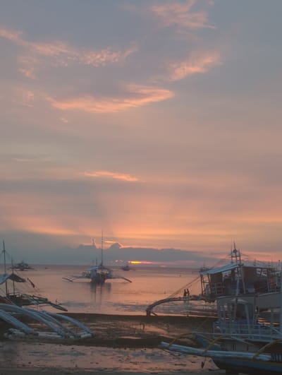 This tranquil image, set in Logon, Philippines, captures a beautiful sunrise or sunset over a coastal area. The scene is dominated by a vast sky, painted with soft, streaky clouds displaying a palette of warm pastel hues—pinks, oranges, and yellows—that gradually blend into cooler blues and grays higher up. The low sun, a vibrant orange glow on the horizon, casts a warm light across the water, which reflects the sky's soft colors.
Several traditional Filipino outrigger boats, known as bangkas, are the primary subjects. In the foreground, a large bangka, featuring a blue and white hull with prominent wooden outriggers, rests partially on a sandy or muddy shore. Further out in the calm water, multiple other bangkas are silhouetted against the radiant sky, their distinct shapes and outriggers clearly visible. The overall impression is one of peaceful serenity, likely at dawn or dusk, as the boats are either docked or anchored, suggesting a quiet moment before or after daily activities. No people or visible text are discernible in the frame. The calm water and soft lighting indicate fair weather.