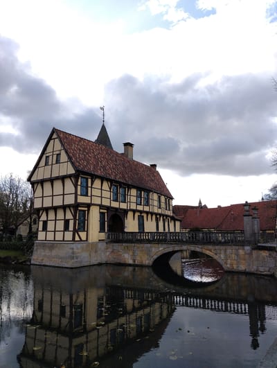 The image displays a historical architectural complex, specifically the Inselhaus (Island House) in Bagno Park, Burgsteinfurt, Germany. A two-story, half-timbered building, characterized by light yellow plaster infill and dark brown exposed timber beams, is situated prominently on the left, directly adjoining a body of dark, still water. The building features a steep, red-tiled gabled roof, from which a slender, conical spire topped with a weathervane extends skyward. Multiple windows with dark blue frames are visible across both floors. An arched stone bridge, constructed from substantial stone blocks and equipped with a balustrade composed of decorative stone posts, spans the water and connects to the building. The entire structure and bridge are reflected distinctly on the water's surface, which also contains scattered green, flat lily pads. To the left of the main building, bare deciduous trees are visible. Additional buildings with similar red-tiled roofs are discernible in the background. The sky is largely overcast with grey clouds, though patches of brighter sky and white clouds are visible towards the upper right. No individuals are present in the frame.