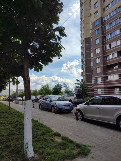 The image depicts a street scene in Bugry, Russia, with a focus on a residential building and parked cars. A multi-story apartment building, constructed with brown and beige bricks, dominates the right side of the frame. The building features numerous windows, some open and some closed, and balconies. In the foreground, a tree with white-painted bark stands on the left side of a paved sidewalk. Several cars are parked along the sidewalk, including a blue sedan, a silver sedan, and a silver SUV. The street is lined with green grass and a few trees. The sky is partly cloudy with white, fluffy cumulus clouds. The overall impression is a typical urban residential area on a bright, possibly summer day.