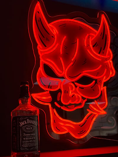A bottle of Jack Daniel's Old No. 7 Tennessee Whiskey stands to the left of a large, red neon sign shaped like a demon's face. The sign is illuminated, casting a red glow on the bottle and the dark background. The demon face has horns, sharp teeth, and stylized eyes. The bottle is clear glass with a black label that features the Jack Daniel's logo and details about the whiskey. The scene is likely indoors, possibly in a bar or a personal collection space, given the neon sign. There is no indication of the city of Campillo Del Moro, Spain in the image.