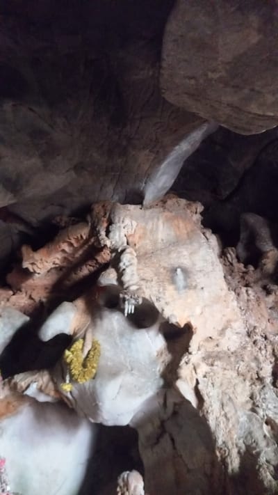 This image shows the interior of Wat Tham Ta Pan temple, a cave temple located in Thap Put, Thailand. The cave walls are made of rock, with a range of crevices and irregular formations. One section has a yellow garland placed on it. Other parts of the rock surface appear to have been carved or decorated, and some areas are layered with stalactites and stalagmites.