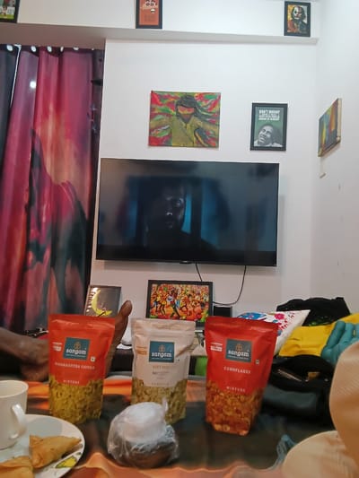 The scene shows a domestic interior, likely in a home in Chopdem, India, with a television mounted on a white wall. The TV screen displays a dark, blurred image of a person, suggesting it might be a movie or show playing.

On the wall around the TV are several framed pictures and paintings, including abstract art and a photograph of Bob Marley. Below the TV, a multicolored curtain is hung.

In the foreground, a glass table holds various items: two red packets of Sangam snack mixtures, including "Maharashtra Chivda" and "Cornflakes Mixture", and a white packet labeled "Diet Mix" with "No Namkeen." There is a plastic bag, a cup, and a plate containing samosas. These items suggest a gathering or snack time is underway.

The background contains additional clothes and objects, giving a sense of lived-in domestic space.