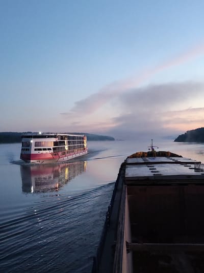The image depicts a tranquil sunrise scene on a wide river. Two vessels are present: a modern, multi-deck passenger ship with a distinctive pink and white hull, and a large, flat-topped barge. The passenger ship is in motion, leaving a wake on the water and its reflection is visible beneath it. The barge, also in motion, is positioned in the foreground, closer to the viewer, and appears to be laden with cargo, though it is partially obscured. The sky is a soft gradient of blue and pink, with wispy clouds illuminated by the rising sun. The riverbanks in the distance are lined with trees and appear to be gently rolling hills. The overall atmosphere is peaceful and serene, capturing a quiet moment on the waterway. The location is Il'inskoe (Lomovskaya S/A), Russia.