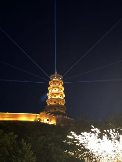 At night, a pagoda-style building is illuminated with yellow light. Blue laser beams extend from the top of the pagoda into the night sky. Adjacent to the pagoda is a structure connected by a bridge-like feature, also illuminated in yellow. A burst of white light, possibly fireworks or sparks, is visible near the top right of the image, close to dark green foliage. The location is likely within the Miyun District, China. There are no people visible in the image.
