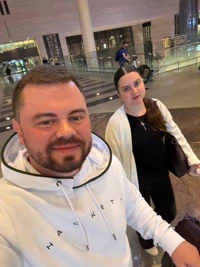A selfie shows a man and woman at an airport. The man in the foreground wears a white hoodie with black letters on it. The woman behind him wears a black top, a white cardigan, and a delicate silver key necklace. Airport architecture is visible in the background, featuring columns and glass walls. Several blurred figures stand or sit in the airport. Luggage and a water bottle are also visible.