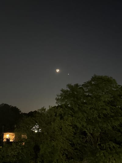 The image depicts a night sky with a crescent moon and a small, reddish celestial body, likely a planet. The moon is illuminated on its right side, and the planet is positioned to the right and slightly below the moon. In the foreground, a large, lush green tree dominates the right side of the frame, its branches swaying. To the left, a house with illuminated windows and a porch light is visible through the trees. The house appears to be nestled among trees on a slope. The overall setting suggests a suburban or residential area at night. The location is identified as Glendale Heights, United States of America.