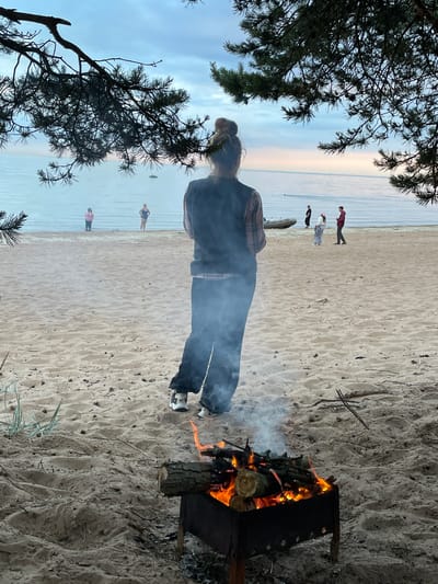 In Lebyazhye, Russia, a bonfire burns on a sandy beach near the Baltic Sea. A young woman stands with her back to the camera, facing the water, with her hair in a bun. Smoke from the bonfire drifts upwards. Several other individuals are visible on the beach and in the water, some appearing to be swimming. A small boat is pulled up on the shore. Pine branches overhang the scene from the upper left, framing the sky and water. The sky is overcast with a soft, diffused light.