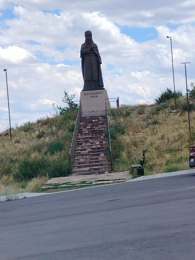 The image shows a statue of a woman dressed in traditional attire, standing on a pedestal. The pedestal has text inscribed on it. A set of brick stairs leads up to the pedestal, and a metal railing runs along the side of the stairs. The statue is situated on a grassy hill with scattered bushes and trees. In the background, there are two tall lampposts and a cloudy sky. To the right of the stairs, a wooden bench with a black trash can is visible. Further to the right, a portion of a vehicle and a wooden fence can be seen. The scene appears to be outdoors, possibly a park or a memorial site, in Qarqaraly, Kazakhstan. The lighting suggests it is daytime. The text on the pedestal reads "ҚАРҚАРАЛЫ АНА".
