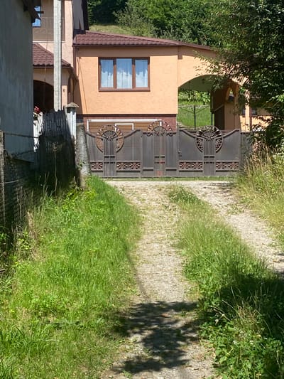 In Luh, Ukraine, a gravel driveway leads to a large, ornate, dark metal gate. The gate is decorated with circular motifs and intricate patterns. Behind the gate, a peach-colored building with a brown tiled roof is visible, featuring a large window with white curtains. The building has an arched entryway to the right. The scene is bordered by lush green grass and foliage, with shadows from trees falling across the driveway. A chain-link fence runs along the left side of the driveway.