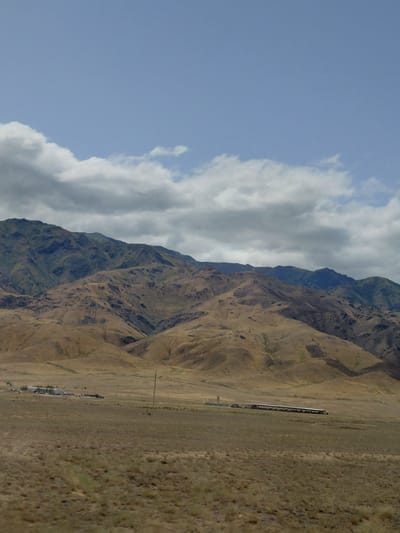 The image depicts a vast, arid landscape with rolling hills and mountains under a partly cloudy sky. In the foreground, a dry, grassy plain stretches out, with scattered vegetation. A lone utility pole stands on the right side of the frame. In the middle ground, a small settlement with several buildings and what appears to be a long, covered structure is visible. There are no people or animals clearly discernible in the scene. The background is dominated by the majestic, textured slopes of mountains, varying in color from golden brown to deep green, suggesting different vegetation or soil types and varying degrees of erosion. The overall impression is one of a remote, natural environment with signs of human habitation. The location is described as Ынталы, Kazakhstan.