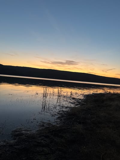The image depicts a serene natural landscape at sunset. The sky transitions from a pale blue at the top to a vibrant orange and yellow hue closer to the horizon, with faint clouds adding texture. A dark, silhouetted hill forms the backdrop, its slope meeting the water. The water's surface reflects the colors of the sky, creating shimmering patterns. The foreground shows the muddy bank of the body of water, covered in dry grass and debris, with some vegetation emerging from the water. Fishing rods are visible on the right side of the bank, suggesting a recreational activity. The scene is likely a rural or semi-rural area near Debelets, Bulgaria, given the natural elements and the presence of fishing gear.