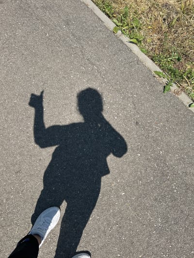 In Dondukovskaya, Russia, a shadow of a person is cast on an asphalt surface, with grass and a concrete curb visible to the right. The shadow depicts the person with their right arm raised and thumb extended upwards, indicating a "thumbs up" gesture. The person's white sneaker is visible in the lower left corner of the frame, alongside dark pants. The scene appears to be outdoors on a sunny day, with the shadow indicating the sun is positioned to the left and slightly behind the photographer. The asphalt is textured and appears to be part of a pathway or road.