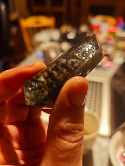 A person is holding a dark-colored, faceted crystal or stone fragment in their fingers. The stone has a polished surface with visible inclusions or variations in color and texture. The background is blurry, but shows a cluttered surface with various containers and objects. A yellow wooden chair is also visible in the background. The location is not possible to determine from the image.