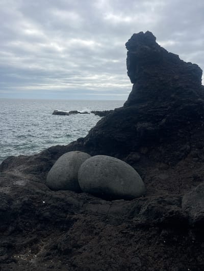 The photograph shows a coastal scene in Lagoa (Nossa Senhora do Rosário), Portugal. Two smooth, gray, round stones are lodged in a dark, rocky cliff face. Above the stones, the rock formation extends outward, forming a natural overhang. Beyond the cliff, the ocean stretches to the horizon under a cloudy sky. The water appears choppy, with a few rocky outcroppings visible in the distance.