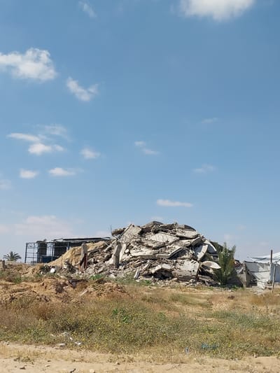 The image depicts a large pile of rubble, likely debris from destroyed buildings, in al-Qarara, Palestinian Territories. The rubble consists of concrete fragments, twisted metal, and other building materials. It sits on a rise of earth, with dry vegetation and a sandy path leading away. In the background, partially obscured by the rubble, are remnants of what appear to be structures with metal frameworks and a few scattered houses with satellite dishes. The sky is clear blue with scattered white clouds.