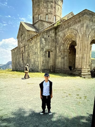 In the city of Artik, Armenia, a young boy wearing a black baseball cap, a white t-shirt, and black pants stands in the foreground on a gravel-covered courtyard. Behind him, the stone facade of a large, ancient building, likely a church or monastery, dominates the background. The building features arched doorways, intricately carved columns, and weathered stone blocks. Two individuals, appearing to be tourists, are walking away from the camera towards the left side of the building. The sky above is a clear blue with scattered white clouds, and the surrounding landscape is hilly with some vegetation. The overall scene suggests a visit to a historical landmark.