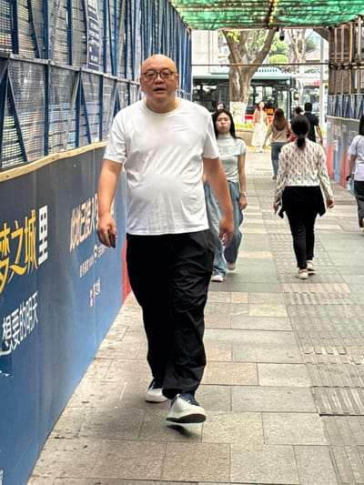 The image shows a man wearing a white t-shirt, black pants, and glasses, standing on a sidewalk next to a blue construction barrier with Chinese writing. In his hand, he holds a dark-colored object, possibly a phone. Behind him, other people walk along the sidewalk, including at least two women in light-colored shirts and jeans. In the background, there are trees and a bus, suggesting an urban setting in 龙溪街道, China. Construction scaffolding is visible on the left side of the frame. The sidewalk is paved with rectangular stone tiles.