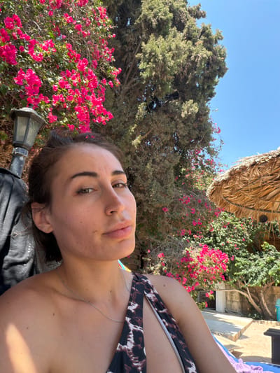 A woman with brown hair and brown eyes is taking a selfie outdoors. She is wearing a black and white patterned swimsuit and a silver necklace. The woman is sitting on a blue lounge chair, and behind her, there are bright pink bougainvillea flowers, a large green tree, and a thatched roof umbrella. The scene appears to be in a sunny, warm climate, consistent with the city of Mazotos, Cyprus. The background includes stone structures and what looks like a pathway.