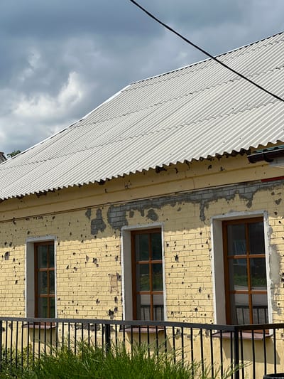 A building in Staryi Saltiv, Ukraine, stands under a cloudy sky. The structure features a white, corrugated roof with a distinct wavy eave. The upper facade is constructed of light yellow brick or painted masonry, displaying numerous small, dark impact marks consistent with projectile damage. Below the roofline, a band of light grey, exposed brickwork is visible where the yellow surface has peeled and flaked. Three rectangular windows with multi-paned, brown wooden frames are set into the damaged facade. In the foreground, a black metal railing with vertical bars extends horizontally, partially obscured by green foliage. A single black utility line traverses the upper right portion of the frame. No individuals are present in the scene.