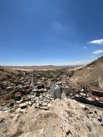 The image shows a view overlooking the city of Ma'loula, Syria, from a high vantage point, possibly a rocky cliff or mountain edge. The sky is clear blue with a few scattered clouds. The terrain is arid and rocky. The city is nestled in a valley and built on the slopes. Buildings are closely packed, mostly low-rise, with a mix of residential and commercial structures. The architecture is traditional, with flat roofs and earth-toned colors. Roads wind through the city. In the background, there are mountains and desert landscape. The overall impression is of a historical settlement in a rugged environment.