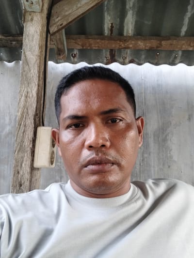 A head-and-shoulders, eye-level, medium shot shows a man looking directly at the camera. The man has dark hair, a light brown complexion, and appears to be sweating. He is wearing a light gray crew-neck t-shirt. To the left of the man, a white electrical outlet is attached to a rough-hewn wooden post. The background consists of a corrugated metal roof above a wall, also made of corrugated metal. The scene appears to be outdoors in a rustic setting, potentially in Sangso, Indonesia.