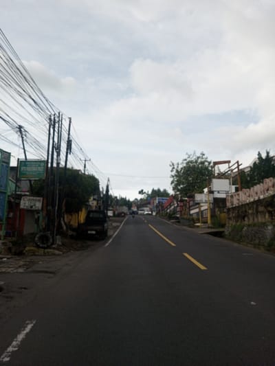 A street stretches into the distance with a center-marked yellow line. Buildings and vegetation line both sides, some with signs visible. Electrical wires crisscross overhead. A dark pickup truck is parked along the left side. The sky is overcast with clouds. The overall scene appears to be a road in a small town or village.