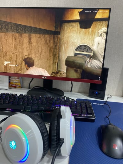 The image shows a gaming setup, including a monitor displaying a video game, a keyboard, a mouse, and headphones. The monitor has a red bezel and displays a scene with a person and architectural elements. Text overlays on the monitor indicate game statistics like frame rate ("кадров/с 184"), GPU usage ("ГП 90%"), CPU usage ("ЦП 68 %"), and latency ("LAT O MC").
Beside the monitor is a small black speaker with blue indicator lights. A keyboard with illuminated keys is positioned in front of the monitor, and a black gaming mouse rests on a blue mousepad with "LORGAR" printed on it. A pair of white gaming headphones with a colorful LED light ring and a logo is placed on top of the keyboard.
The scene appears to be an indoor setting, likely a desk area for gaming or computer use. The overall impression is of a modern gaming or computer setup, with attention to visual aesthetics, as suggested by the RGB lighting on the headphones and keyboard. The context of Achinsk, Russia, does not provide specific visual cues in the image itself, but it grounds the location of the depicted scene. There is no indication of the time of day or weather.