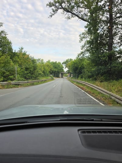 The image captures a view from the driver's perspective inside a vehicle, looking forward on a paved road. The road is bordered by a metal guardrail on both sides and lined with lush green trees. In the distance, an overpass with concrete pillars is visible, suggesting the road passes under a bridge. There are several traffic signs near the overpass. The sky above is partly cloudy with a light blue hue. The foreground shows the dashboard of the car, with a speaker grille visible. The scene appears to be a road on the outskirts of Kelsterbach, Germany, characterized by its natural, tree-lined surroundings and infrastructure elements like the road and overpass. There are no people visible in the image.