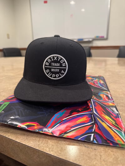 A close-up, rotated shot shows a black snapback hat with a circular logo on the front. The logo is white and embroidered with the words "BRIXTON TRADE MARK SUPPLY" around the circumference, and a "B" in the center. The hat is placed on top of a colorful, abstract printed surface. The surface appears to be paper or cardstock with a dynamic design of broad brushstrokes in red, yellow, blue, and orange against a black background. This printed material is partially folded. In the background, a whiteboard is visible, with some faint markers or stains on it. A wooden table surface fills the lower part of the frame and extends into the background. The overall setting appears to be an indoor space, possibly an office or a room with a whiteboard. There is no indication of the time of day or weather. The composition is dominated by the hat and the vibrant printed surface.