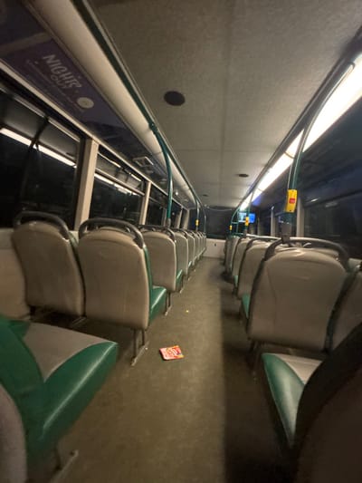 The interior of a bus is depicted with rows of seats. The seats have light-colored backs and green headrests. Handrails run along the bus's length above the windows. A "NIGHTS OUT" advertisement is visible on the lower wall by the windows. The bus's interior is dimly lit with overhead lighting. A small, rectangular object rests on the floor between the rows of seats. The perspective is slightly tilted.