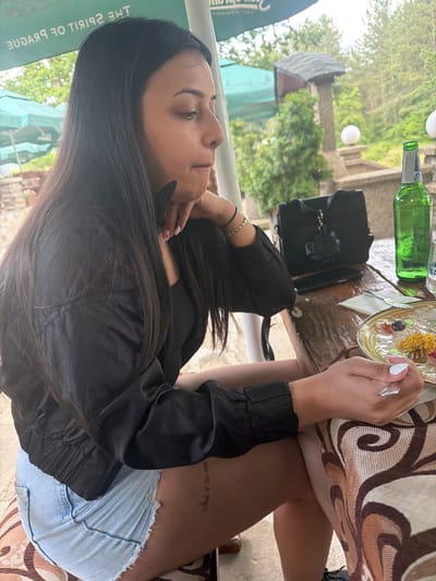 An eye-level outdoor shot captures a woman seated at a table, seemingly enjoying a meal in Sushina, Bulgaria. The image is taken from a slightly elevated perspective, looking down and across. The woman, with long dark hair, is wearing a black jacket and light denim shorts. Her right hand, adorned with white nail polish, holds a fork above a clear plate containing what appears to be oatmeal or a similar dish with berries.

On the wooden table, a patterned tablecloth with brown and beige swirls is visible. To her left, a green glass bottle, possibly beer, and a black handbag are placed. Her left hand rests near her chin, suggesting a moment of thought or relaxation. A small tattoo with illegible text is visible on her upper left thigh/hip area.

The background reveals a lush green outdoor setting with numerous trees and foliage. White spherical lights are interspersed among the greenery. Part of a green banner or awning is visible on the left, displaying reversed text that reads "THE SPIRIT OF PRAGUE". The overall ambiance suggests a casual, pleasant daytime meal at an outdoor cafe or restaurant.