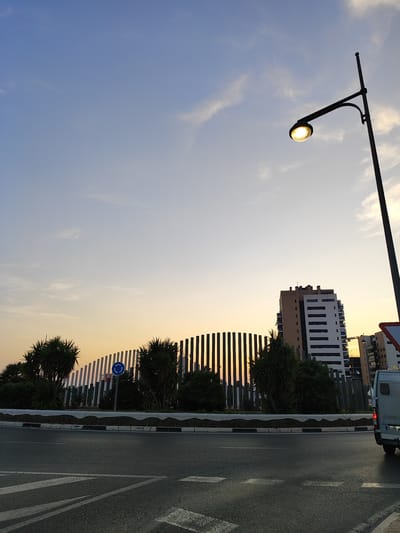 The image captures a street-level view of El Campello, Spain, at dusk or dawn. A street lamp with a lit bulb illuminates the scene from the upper right. In the background, a modern white and beige building with multiple balconies stands tall, flanked by a row of vertical, dark metal structures that form a curved fence or wall. Palm trees are visible in front of this fence, adding a touch of greenery. A traffic sign, partially visible on the right, appears to be a warning sign. The foreground shows a paved road with white lane markings, indicating it is a street or intersection. A portion of a white van is seen on the far right side of the image, suggesting it is parked or driving by. The sky above transitions from a pale blue at the top to a warm orange and yellow hue near the horizon, characteristic of a sunrise or sunset. The overall atmosphere is calm and urban, with a hint of natural light.