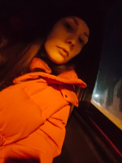 A person with light skin and dark hair is illuminated with a yellow glow in a dark setting. They are wearing a puffy, bright orange jacket with visible seams and a button detail. A car window with a blurry reflection is visible to the right, suggesting the individual is inside a vehicle. The image is taken at an angle, focusing on the person's face and the jacket.