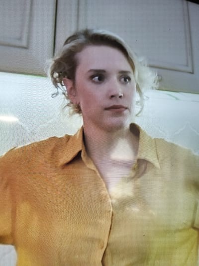 The image is a medium shot of a young woman with blonde, messy hair and fair skin. She is wearing a yellow collared shirt. Her eyes are wide and looking to her left, and her mouth is slightly open. The background appears to be an interior setting with light-colored cabinets or walls. There is no visible indication of the city of Sloboda, Russia, or any specific location. The image quality is somewhat grainy, suggesting it might be a still from a video or a lower-resolution photograph.