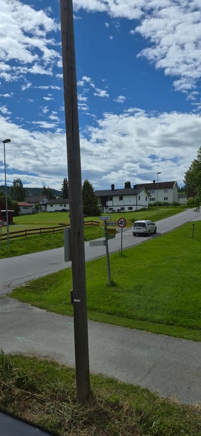 This media file shows a daytime scene in Biri, Norway, with a bright blue sky filled with white, fluffy clouds. The perspective is tilted, giving a sense of looking up or from a slightly angled viewpoint.

The scene features a paved path winding through a grassy area with several residential buildings. The houses appear to be of modern design, with light-colored facades and dark roofs. A white car is parked by the side of the road, near a street sign and a speed limit sign displaying "40". Another, larger wooden beam or pole stretches horizontally across the foreground.

In the distance, a red fence lines part of the property. The overall impression is of a peaceful, residential setting on a clear, sunny day. The sky suggests it is late morning or early afternoon. There is no visible text on the car, houses, or fence. The street signs have text, but it is not clearly legible in this resolution.